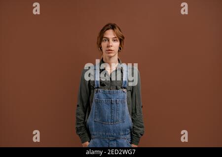 Photo of cute woman farmer stands with serious face. Female wears denim overalls, isolated brown color background Stock Photo