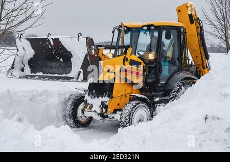 Yellow tractor excavator with a large bucket clears a snowy road from snow Stock Photo