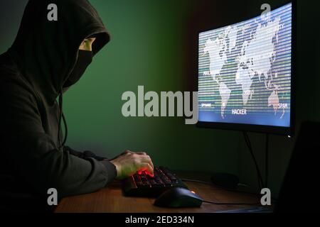 Masked hacker typing in a computer for organizing massive data breach attack around the world. Cyber criminal concept. Stock Photo