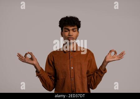 Photo of black man looks relaxed and have meditation. Male wears shirt, isolated grey color background Stock Photo