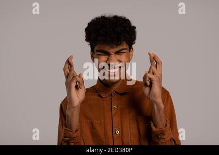 Photo of black man crosses fingers and looks worried about something. Male wears shirt, isolated grey color background Stock Photo