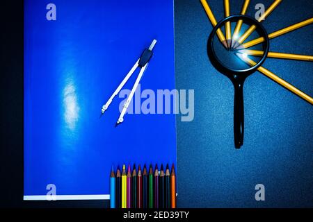 On top of a large blue notebook is placed a compass, on the side a magnifying glass with pencils, at the bottom of the crayons. Stock Photo
