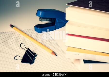 A stapler with a pencil and a clip are placed on a checked sheet, in the foreground a stack of books. Stock Photo