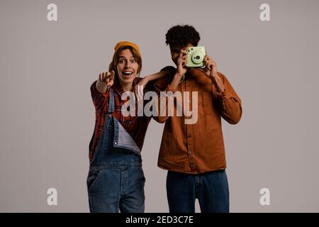 Photo of cute couple farmers take a photo on polaroid and looks happy together . White woman wears denim overall, black man shirt isolated grey color Stock Photo