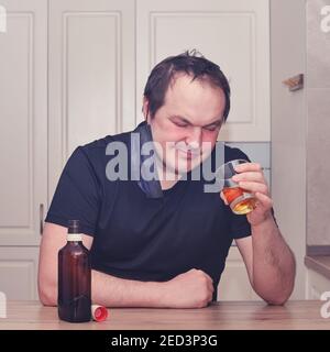 A man looks thoughtfully at a glass of whiskey sitting in a home kitchen. Problems with drinking alcohol during a pandemic, isolation and lockdown Stock Photo