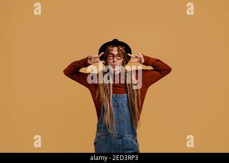 Photo of attractive female farmer looks happy, wears funny glasses. White woman wears denim overall and hat isolated brown color background Stock Photo