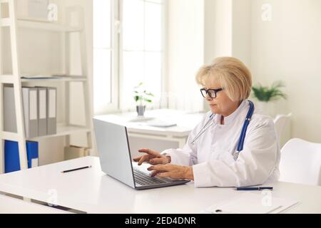 Mature woman doctor in white uniform sitting and working online on laptop Stock Photo