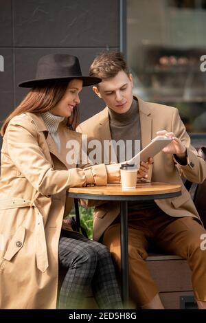 Young restful couple sitting by small round table against modern architecture, having drinks and discussing online news while using tablet Stock Photo