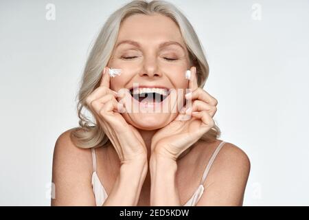 Happy funny 50s middle aged woman applying cream on face isolated on white. Stock Photo