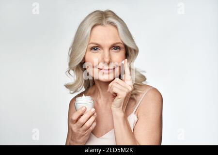 Smiling 50s middle aged woman putting facial cream on face looking at camera. Stock Photo