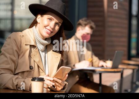 Young elegant brunette businesswoman in hat, sweater and trenchcoat using smartphone and looking at you against man networking Stock Photo