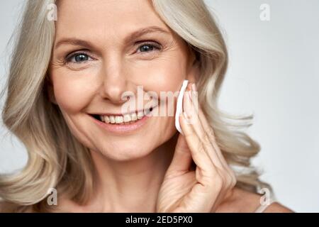 Smiling 50s middle aged mature woman holding cotton pad cleaning face skin. Stock Photo