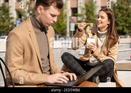 Young serious elegant man using laptop while sitting on bench in urban environment on background of his happy girlfriend eating wok Stock Photo