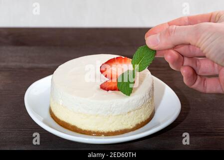 Cheesecake cake with fresh strawberries on white plate. Woman's hand decorated cake a mint. Stock Photo