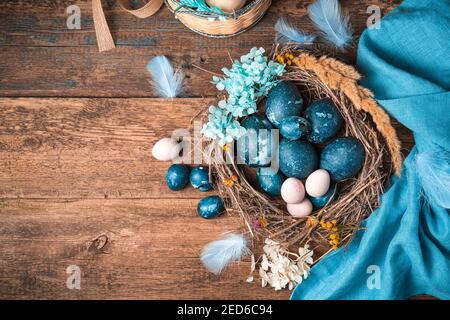 A set of quail and chicken eggs in dark blue and pastel colors in a beautiful nest with flowers and feathers. Stock Photo