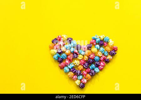 Colored popcorn. Heart shape made of colored popcorn, flat lay on yellow background with copy space, Stock Photo