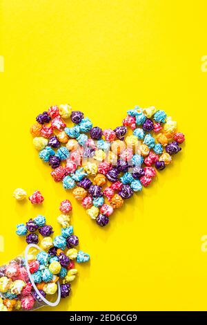 Colored popcorn. Heart shape made of colored popcorn, flat lay on yellow background with copy space, Stock Photo