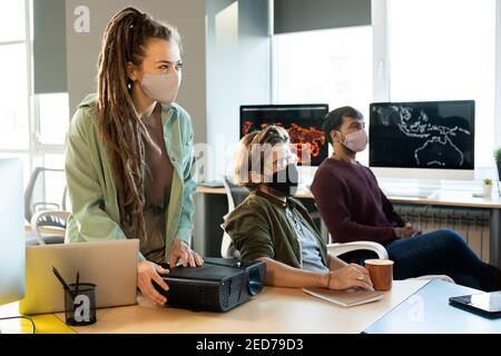 Young businesswoman or coach in casualwear and protective mask using projector while making presentation for colleagues at seminar Stock Photo