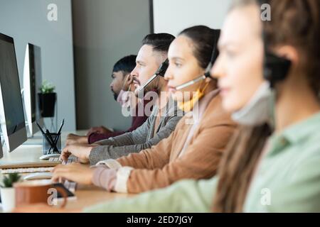 Young serious male operator with protective mask under chin consulting clients online while sitting among colleagues in modern call center Stock Photo