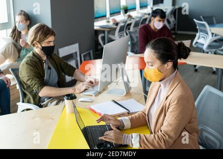 Group of young co-workers in casualwear and protective masks sitting in open space office and using mobile gadgets during individual work Stock Photo