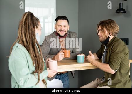 Two young confident businessmen and their female colleague with mugs having coffee by table in office during conversation at break Stock Photo