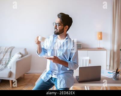 Eastern Man Relaxing With Cup Of Coffee After Working At Home Office Stock Photo