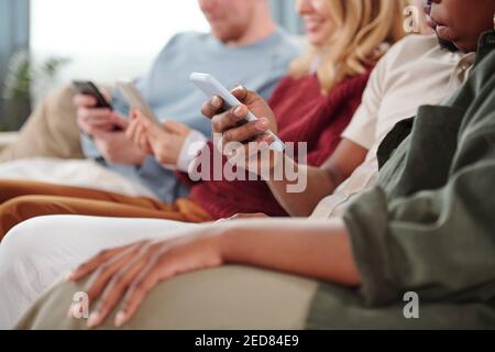 Group of young contemporary intercultural friends sitting in row on couch and scrolling in their mobile gadgets, focus on African couple Stock Photo