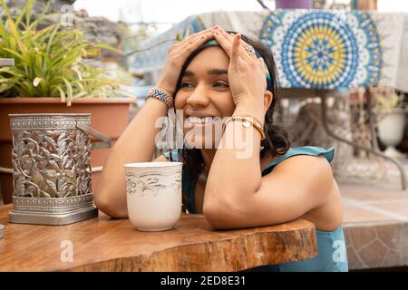 cheerful young woman with brown skin sitting at a wooden table, smiling next to a cup of coffee outside, on a sunny day on vacation Stock Photo