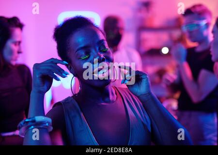 Happy young glamorous woman of African ethnicity in smart casualwear keeping eyes closed while standing in front of her dancing friends Stock Photo