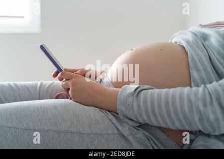 Side view on midsection of pregnant caucasian woman and belly lying on the bed with bed with mobile phone reading internet or sending message calling Stock Photo