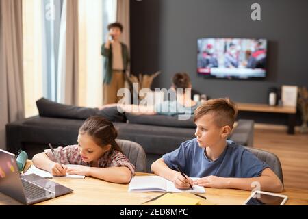 Two contemporary schoolkids sitting by table in front of laptop, making notes in copybooks and listening to their teacher during online lesson Stock Photo