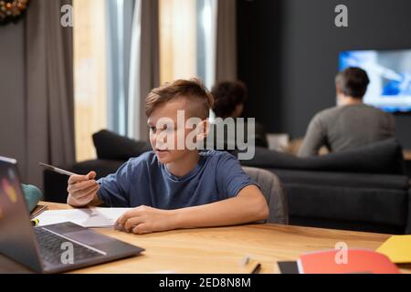 Cute and clever schoolboy with pen looking attentively at laptop display while sitting by table and listening to his teacher during online lesson Stock Photo
