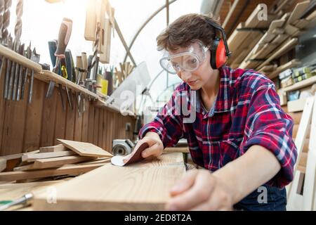Young attractive 30-40 professional female carpenter looking grinding raw wood with sandpaper in carpentry diy workshop. Feminine women equality Stock Photo