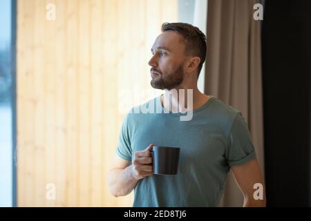 Young bearded relaxed man in grey t-shirt holding mug with hot tea or coffee and looking through large window while staying at home Stock Photo
