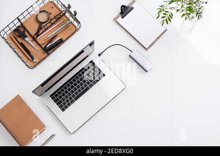 Top view of white desk with laptop, basket with office supplies, blank papers in clipboard, green plant in glass, notebook and silver pen Stock Photo