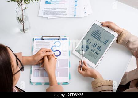 Two young businesswomen analyzing and discussing graph, charts and diagrams while one of them pointing at tablet screen at meeting Stock Photo