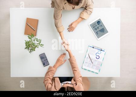View of two young brunette businesswomen shaking hands over desk after making deal among financial document, notebook and mobile gadgets Stock Photo