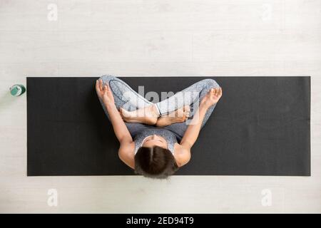 View of young brunette sportswoman in tracksuit keeping legs crossed while sitting on mat in pose of lotus, relaxing or meditating Stock Photo