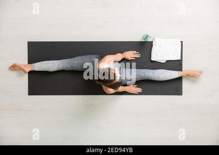 View of young active brunette female in tracksuit stretching legs in front of and behind herself while exercising alone on mat during training Stock Photo