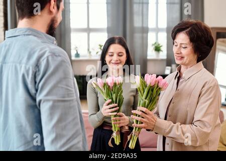 Two happy brunette females in smart casualwear looking at bunches of fresh pink tulips in their hands while standing in front of young man Stock Photo