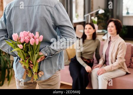 Rear view of young man in casualwear with two bunches of fresh pink tulips behind his back standing in front of two happy females Stock Photo