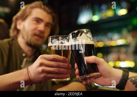 Hands of two friendly guys clinking with glasses of beer in front of camera while one of them pronouncing toast against bar with alcoholic drinks Stock Photo