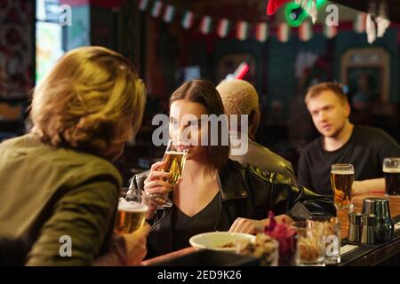 Young brunette female in black leather jacket and pullover drinking beer and looking at her boyfriend during chat by bar counter against two guys Stock Photo
