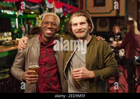 Two young cheerful intercultural men looking at you while toasting with glasses of beer in front of camera against guests sitting by table in pub Stock Photo