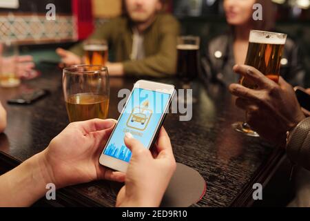 Hand of young woman with smartphone over table going to call for taxi against group of friends having drinks and discussing their plans Stock Photo