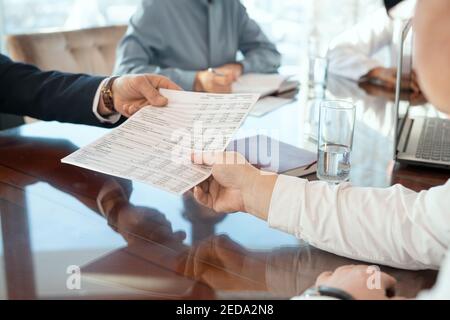 Hand of mature male delegate taking document with financial data being given by business partner against secretary making notes in notebook Stock Photo