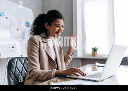 Beautiful business woman communicates by video conference. Young adult female employee is sitting at the desk in office, using laptop and smiling. African american woman distantly working or studying Stock Photo