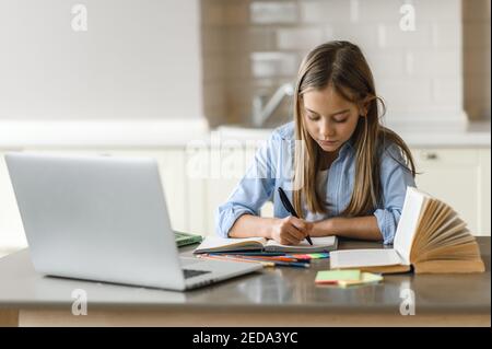 Distance learning, homeschooling during quarantine. Caucasian pretty schoolgirl doing her homework at home using laptop, sitting at the work desk Stock Photo