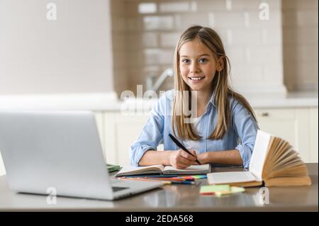Caucasian pretty blonde schoolgirl is learning at home using laptop, sitting at the work desk, looks at the camera and smiling. Education during quarantine Stock Photo