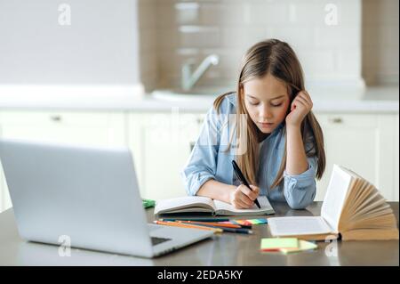 Distance learning, homeschooling during quarantine. Focused caucasian pretty schoolgirl doing her homework at home using laptop, sitting at the work desk Stock Photo